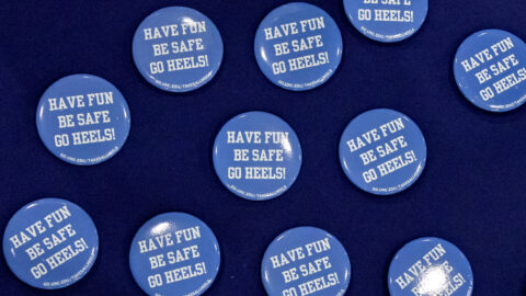 Carolina blue buttons displayed on a navy blue table cloth. Buttons say, "HAVE FUN, BE SAFE, GO HEELS!" in white.