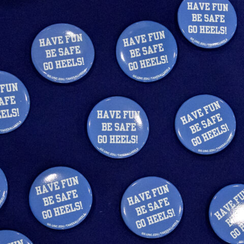 Carolina blue buttons displayed on a navy blue table cloth. Buttons say, "HAVE FUN, BE SAFE, GO HEELS!" in white.
